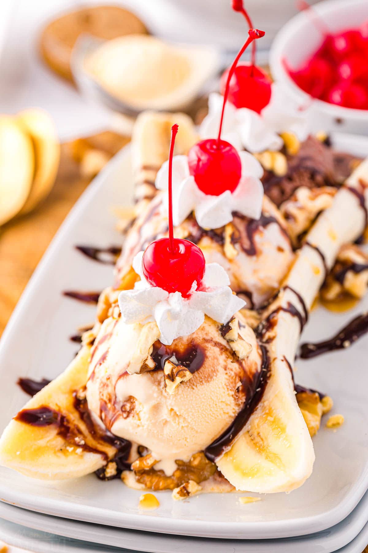 front view of an impressive banana split made on a white rectangular plate. Topped with all the classic toppings and bright red maraschino cherries.