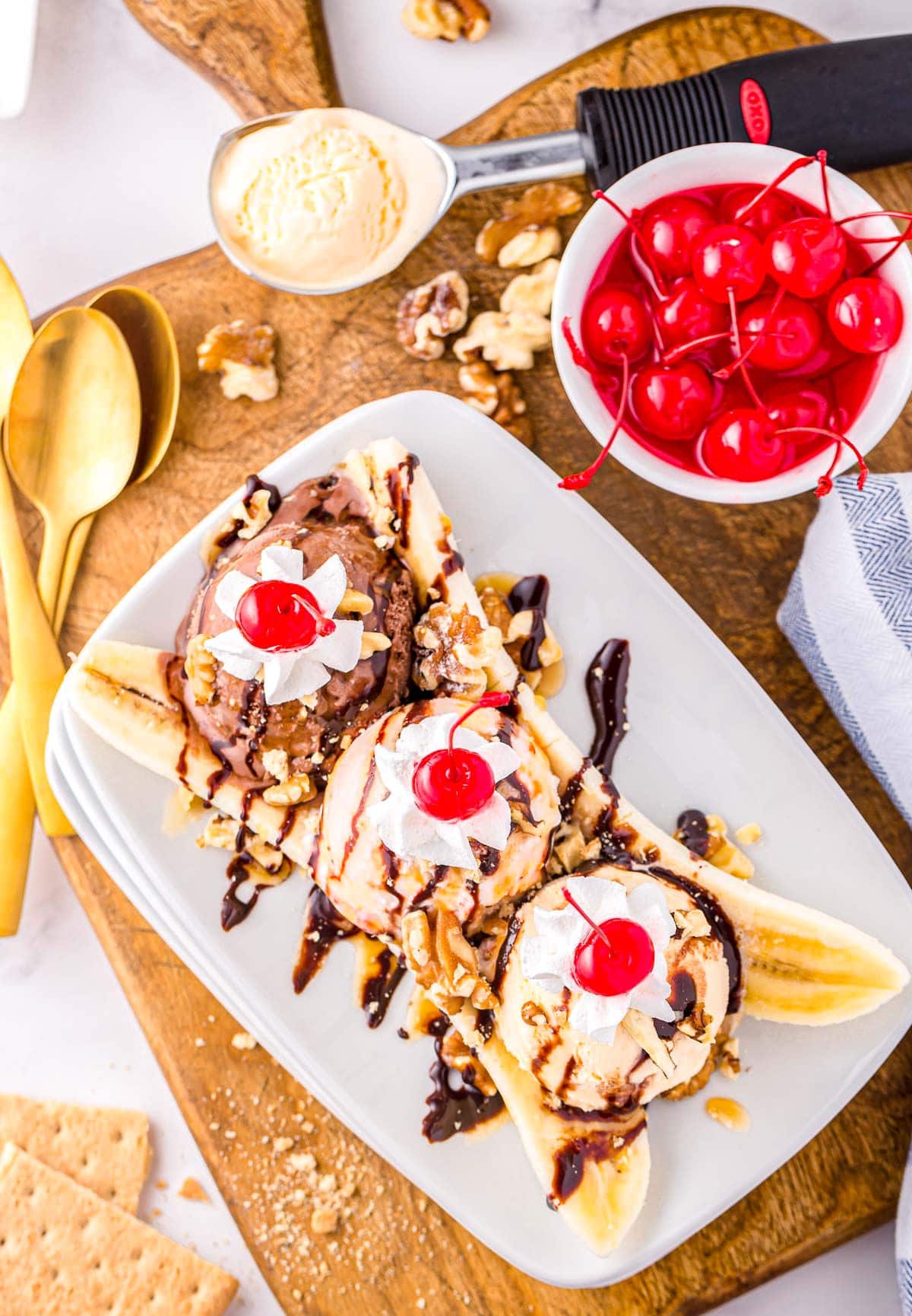 top down look at a classic banana split made with three different flavors of ice cream, whipped cream, and cherries. Plate is sitting on wood board next to a small bowl of maraschino cherries.