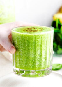 hand holding green smoothie in ridged glass with greens in the background.