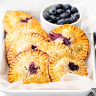 7 blueberry hand pies in a parchment lined sheet pan with fresh blueberries in a bowl in the back.