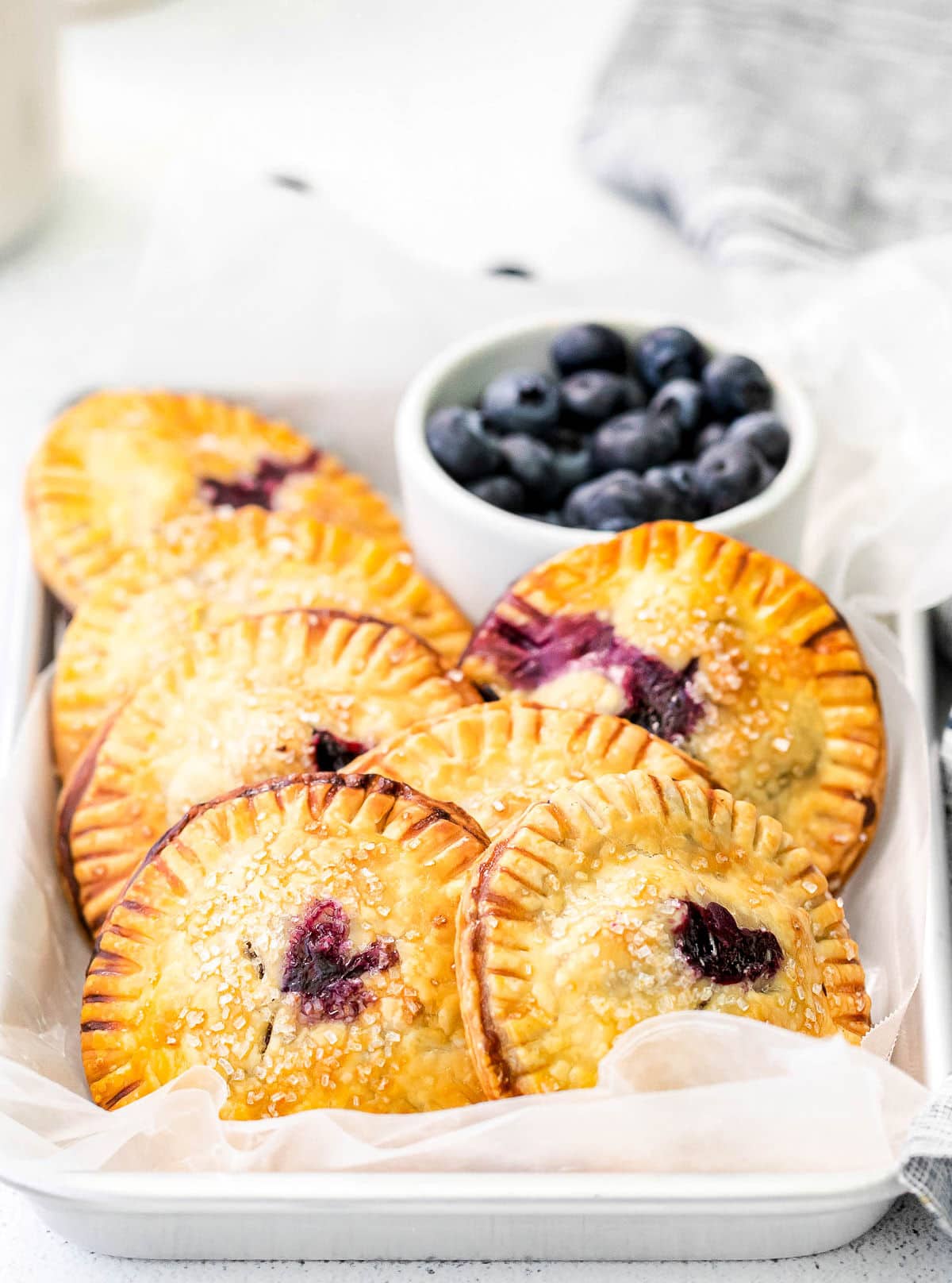 7 blueberry hand pies in a parchment lined sheet pan with fresh blueberries in a bowl in the back.