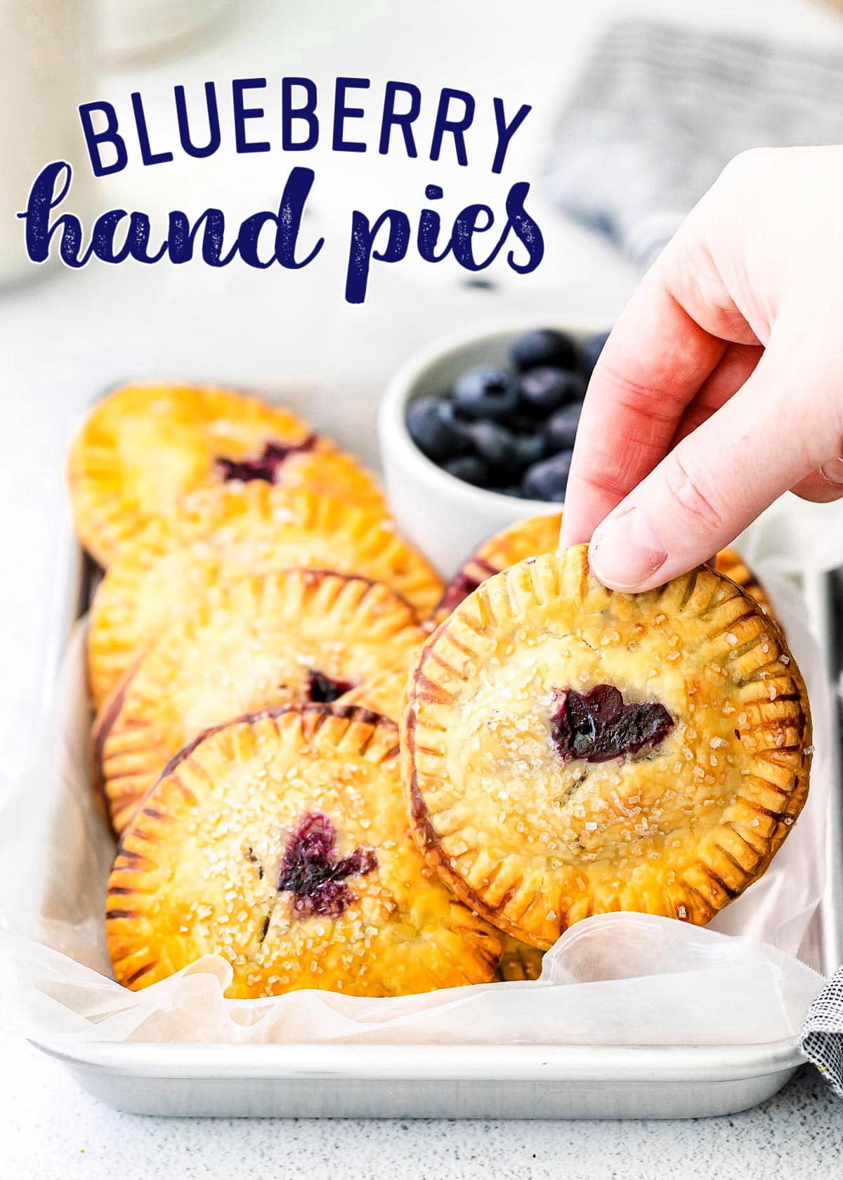 six blueberry hand pies on a quarter sheet tray with a small bowl of blueberries. One pie is being held up with a hand. Title overlay at top of image.