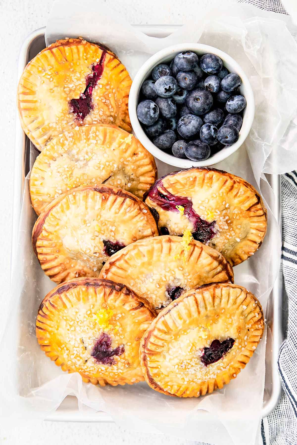 top down look at blueberry hand pies ready serve from small sheet pan with small bowl of blueberries nestled next to the pies.