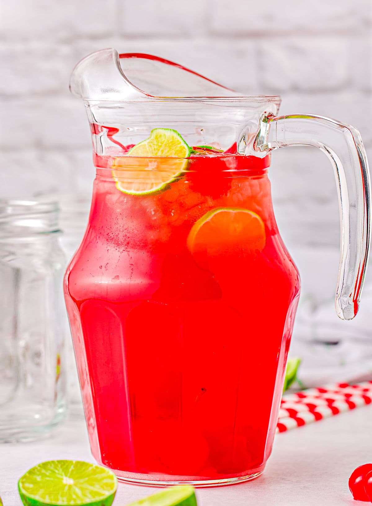 cherry limeade in a tall glass pitcher with fresh lime slices in and around the pitcher as well.