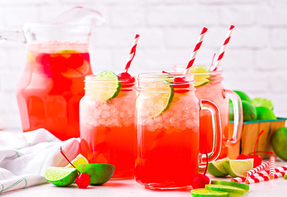 three mason jars plus a tall pitcher filled with cherry limeade and garnished with lime slices and maraschino cherries.