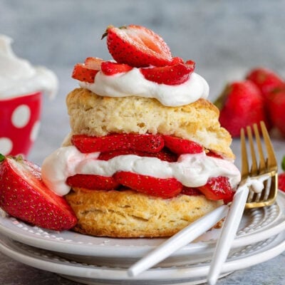 strawberry shortcake on white plate with layers of macerated strawberries and freshly whipped cream. two forks resting on the edge of the plate on one side and half a strawberry on the other.