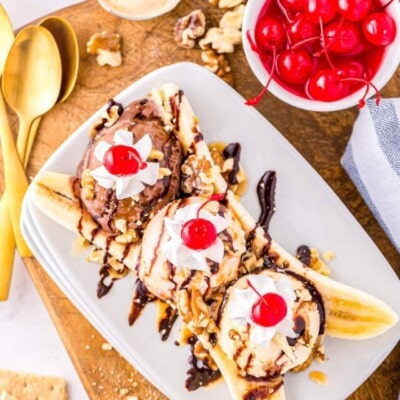top down look at a classic banana split made with three different flavors of ice cream, whipped cream, and cherries. Plate is sitting on wood board next to a small bowl of maraschino cherries.