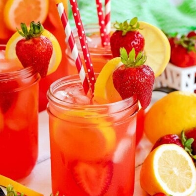 three mason jars filled with strawberry lemonade and topped with lemon slices and fresh strawberries. Red and white paper straws in jars.