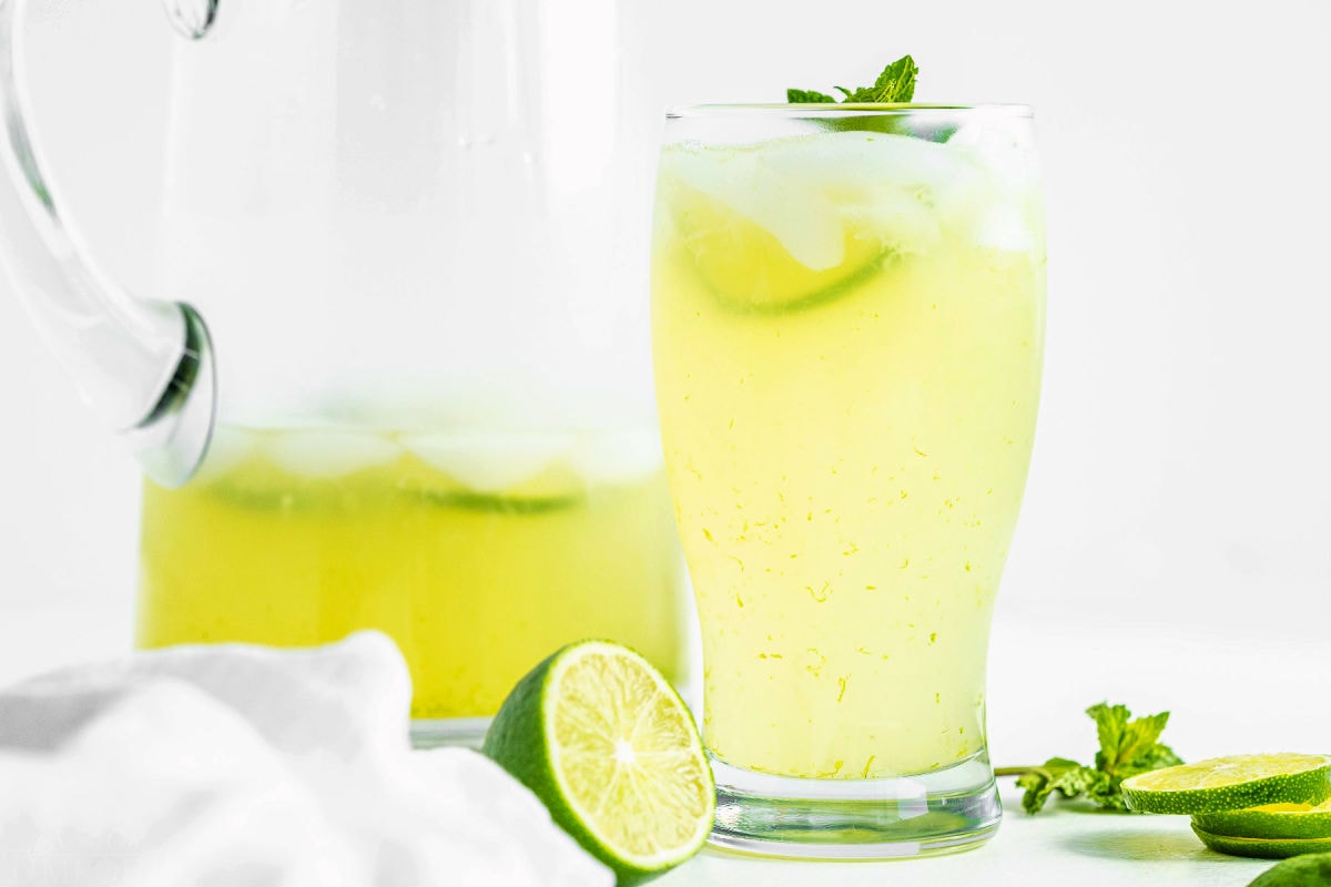 wide look at glass of limeade sitting next to a half full glass pitcher with more limeade in it.