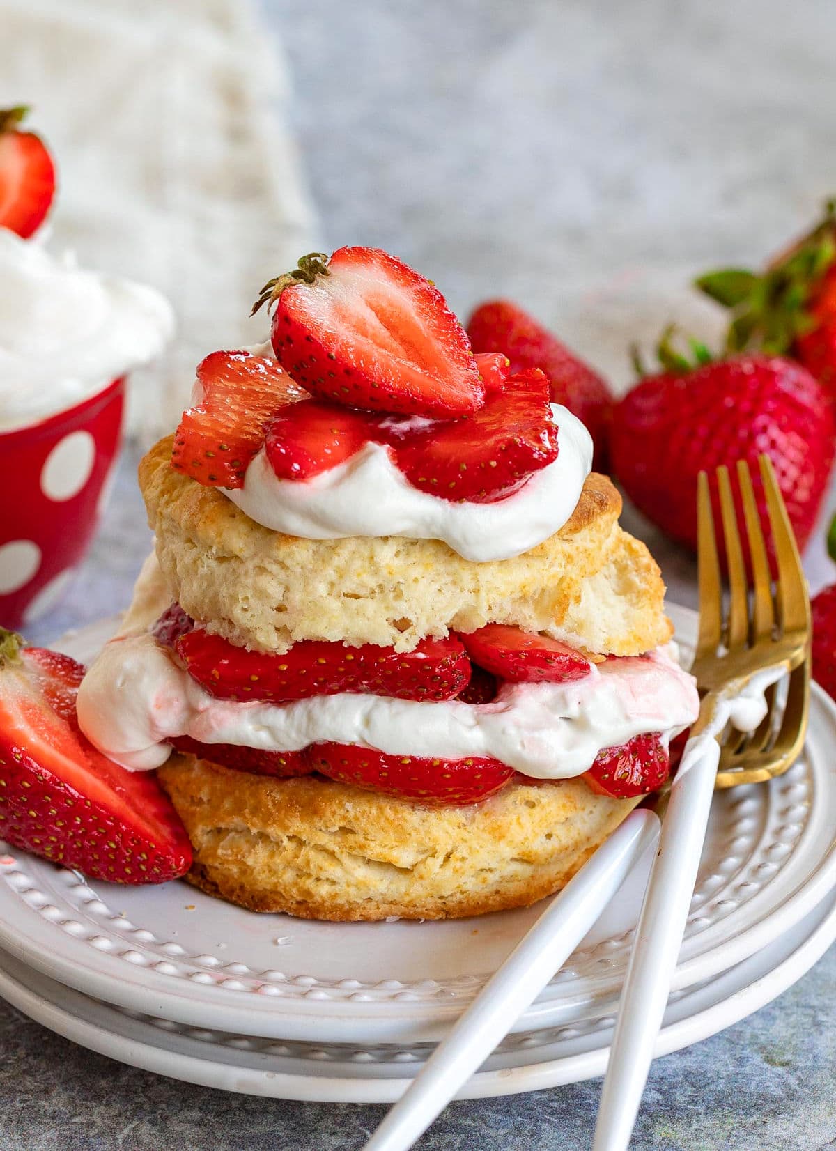 strawberry shortcake on white plate with layers of macerated strawberries and freshly whipped cream. two forks resting on the edge of the plate on one side and half a strawberry on the other. beige napkin in background.