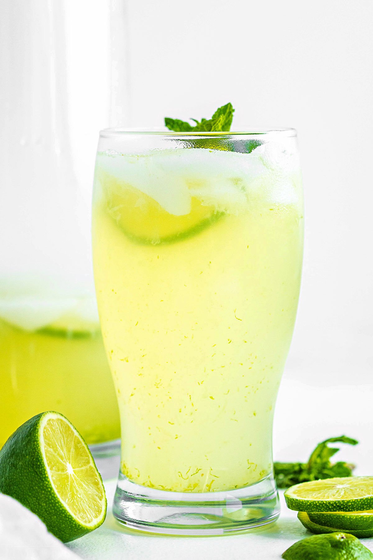 tall glass of freshly made limeade garnished with a sprig of mint and fresh lime slices sitting in front of pitcher of limemade.