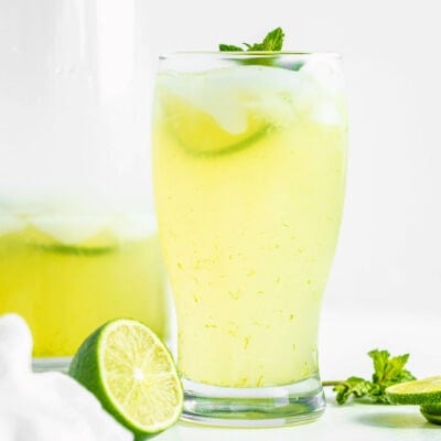 glass of limeade sitting in front of tall glass pitcher with more limeade in it. Fresh lime slices and mint sprigs scattered about.