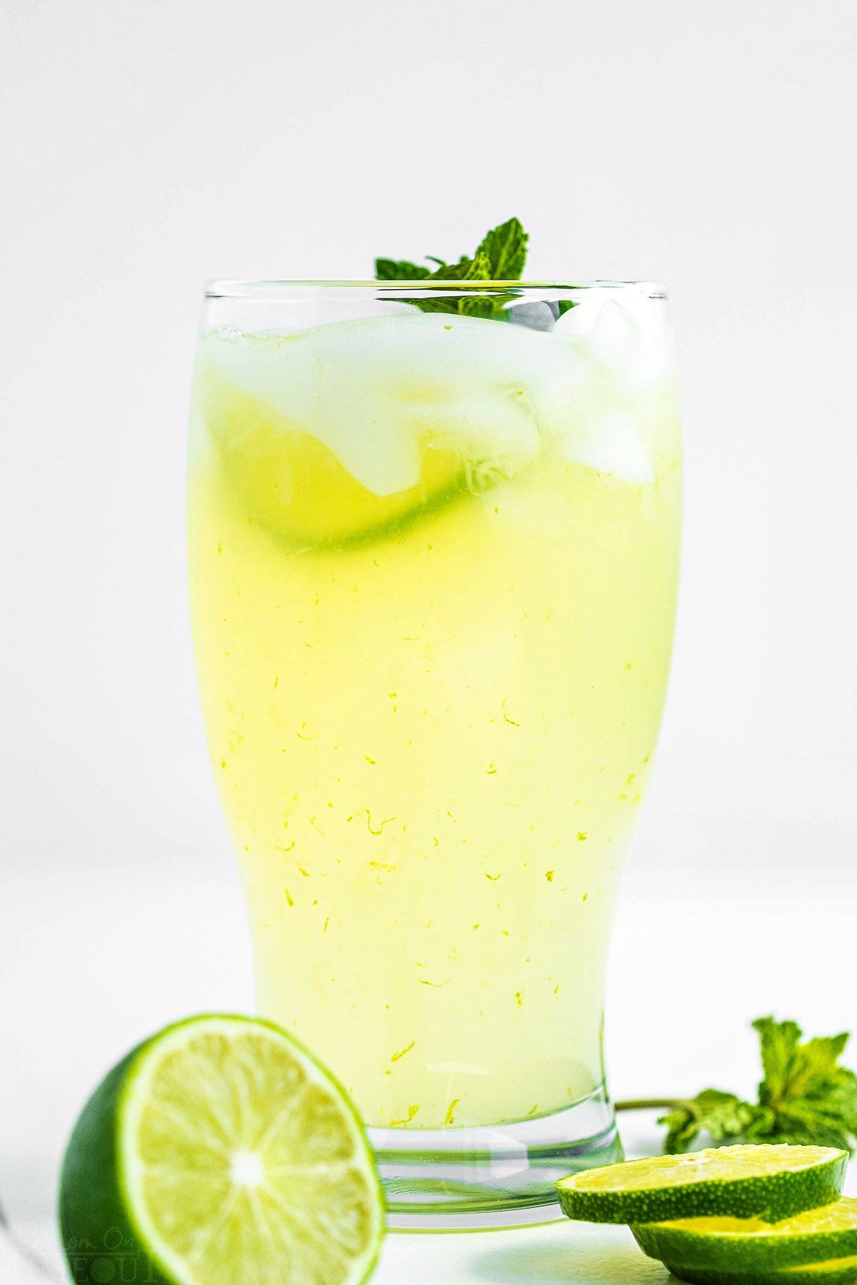 tall glass of freshly made limeade garnished with a sprig of mint and fresh lime slices sitting in front of pitcher of limeade.