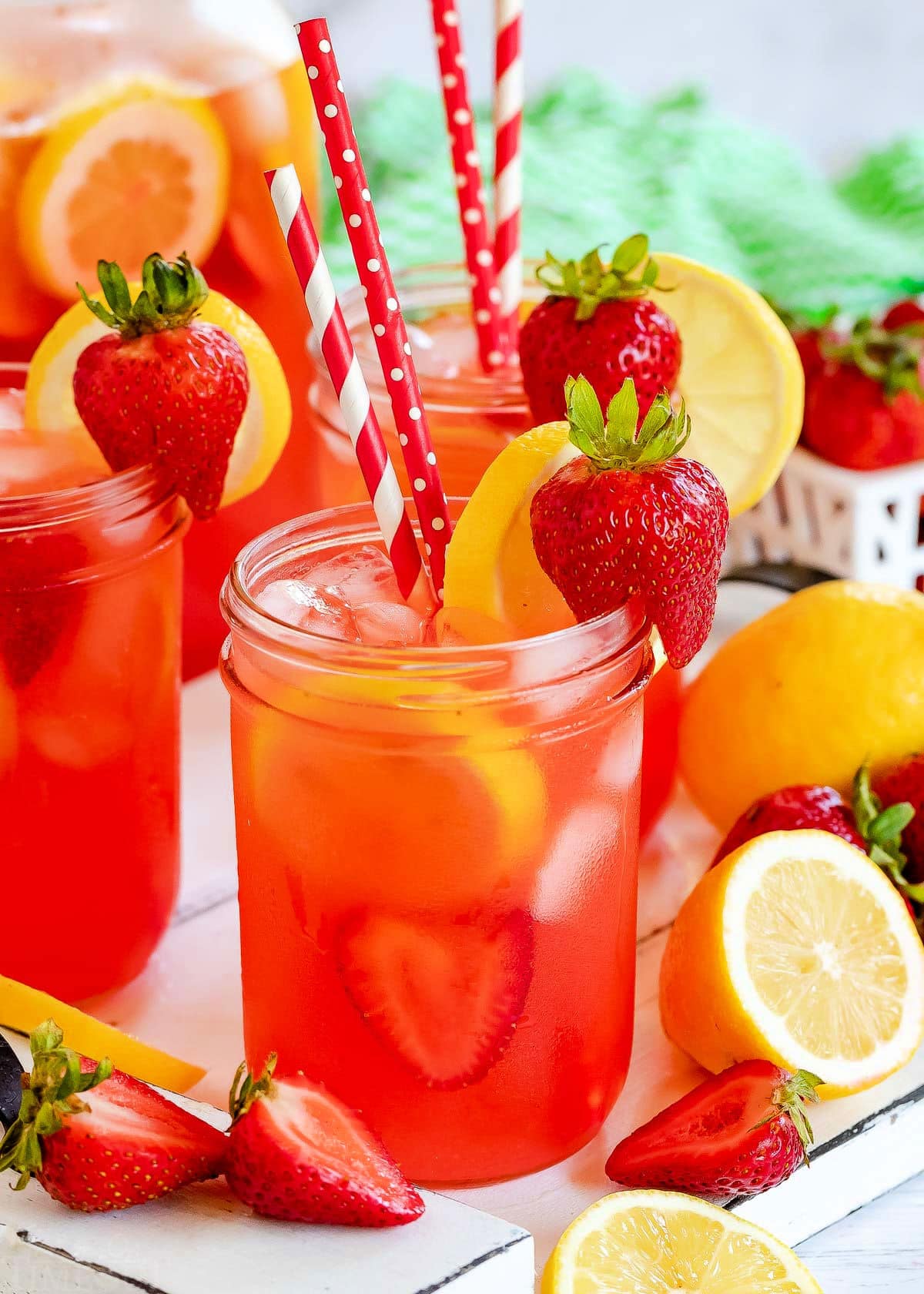 three mason jars filled with strawberry lemonade and topped with lemon slices and fresh strawberries. Red and white paper straws in jars.