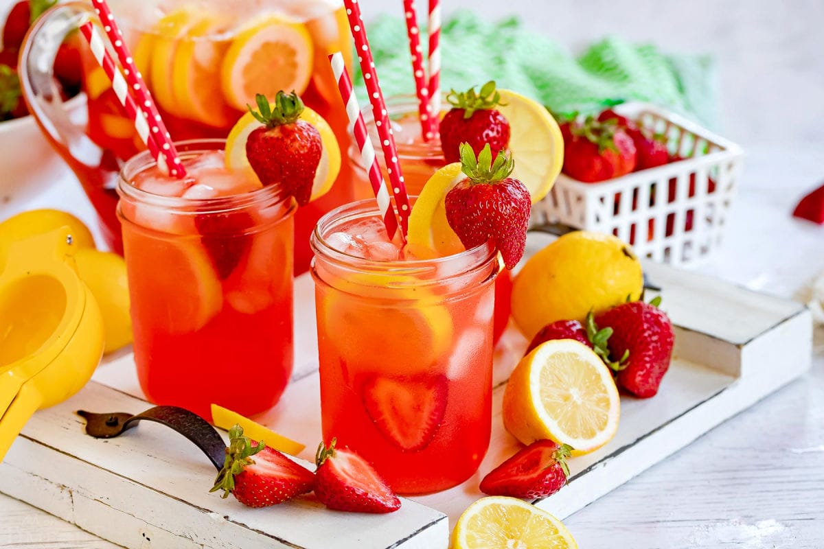 three mason jars filled with strawberry lemonade and topped with lemon slices and fresh strawberries. Red and white paper straws in jars.