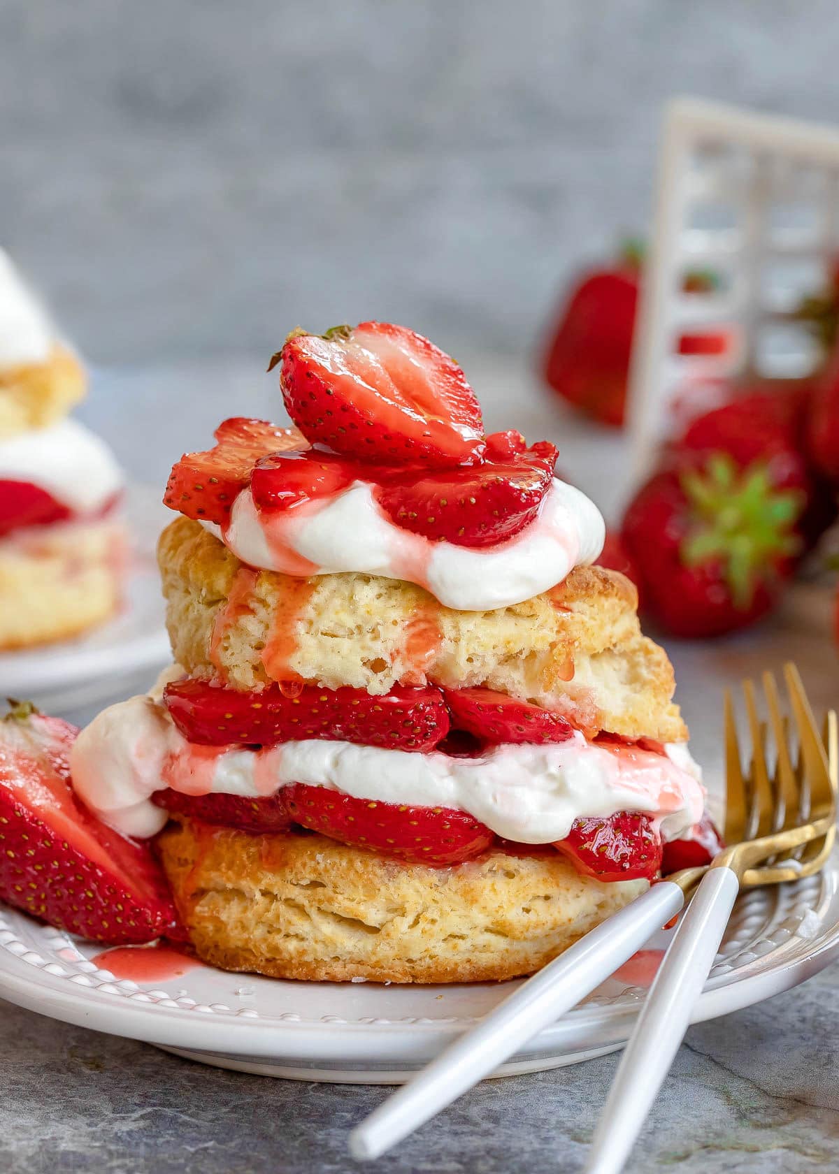 strawberry shortcake made with biscuits and macerated strawberries and fresh whipped cream. Layered on a white plate with two forks sitting on the edge of the plate.