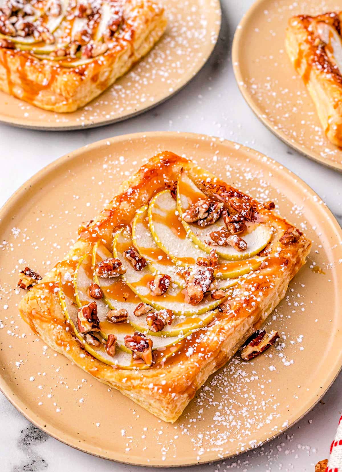 single apple tart on light orange plate with two more apple tarts in the background.