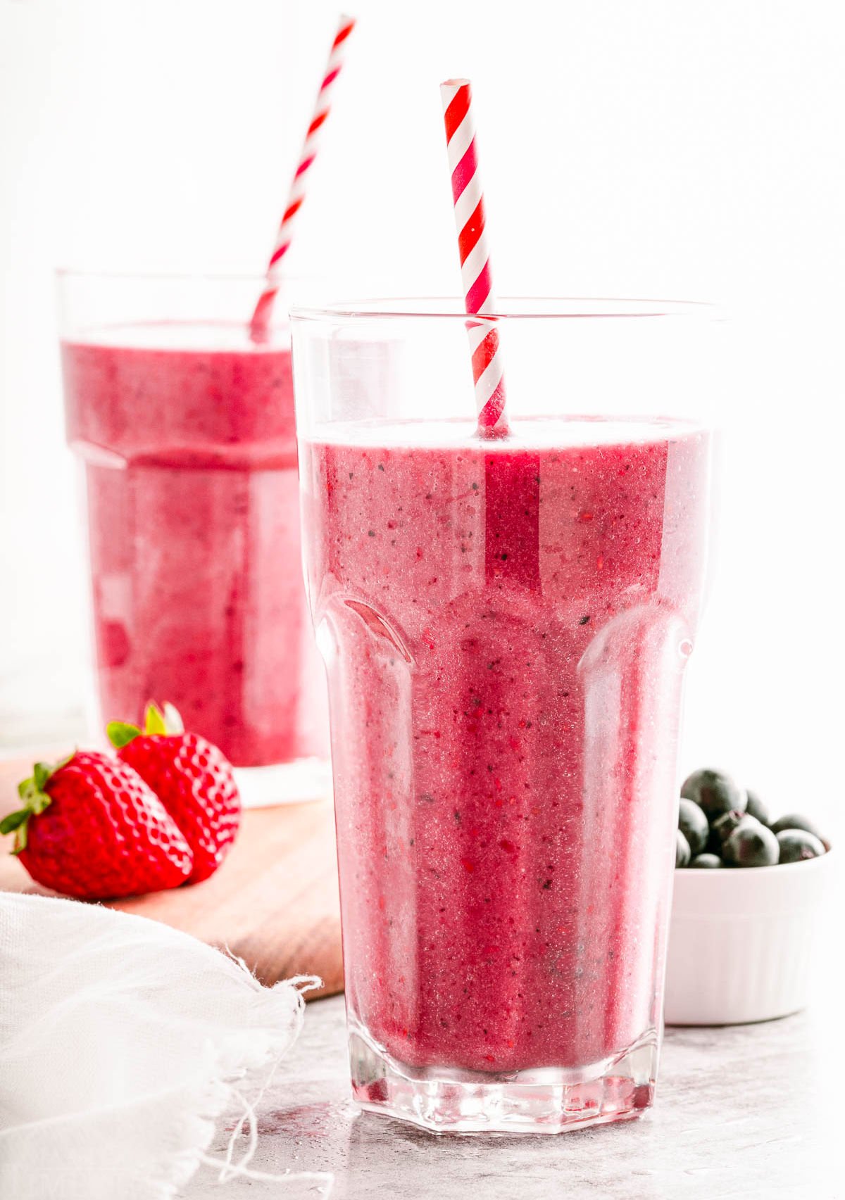 two glasses filled with berry smoothies sitting on and next to a small wood board. both smoothies have striped paper straws in them.