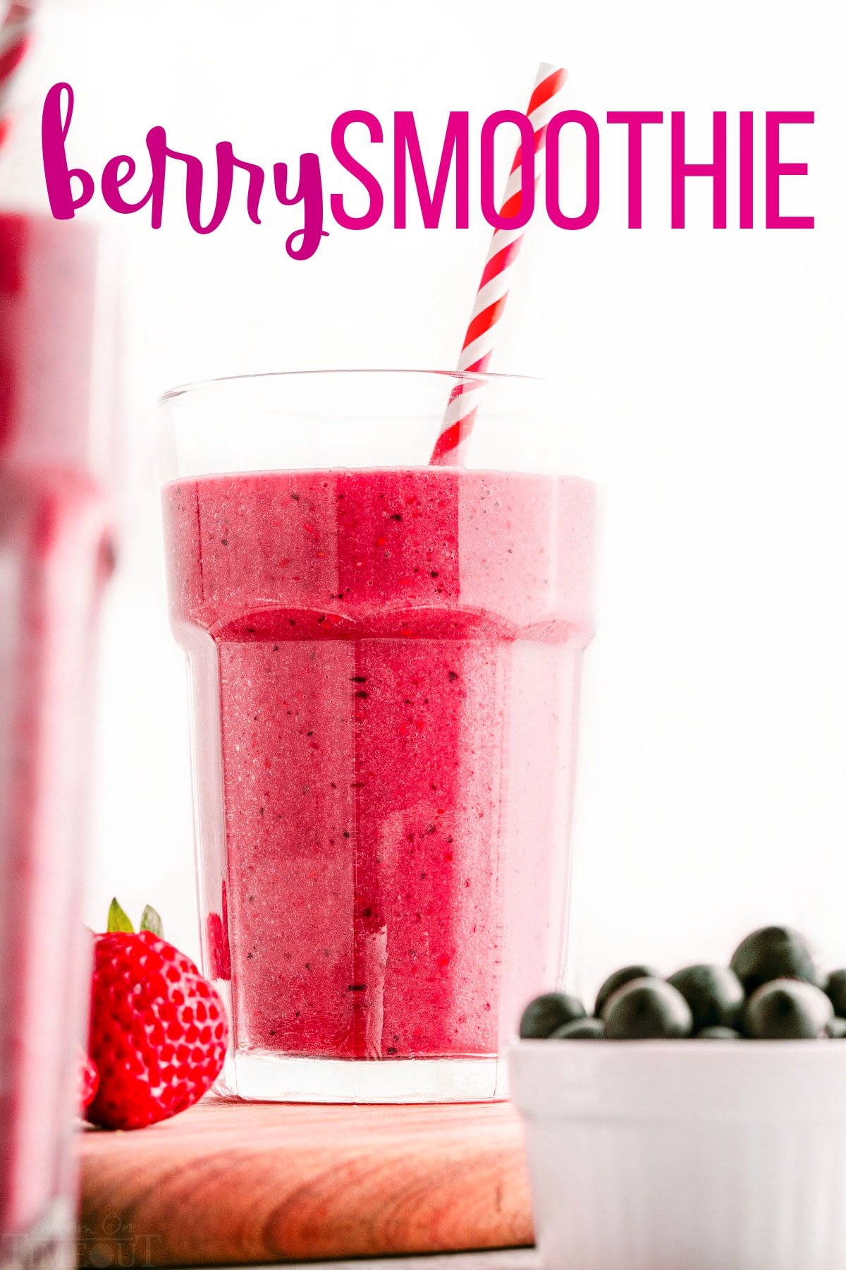 berry smoothie in tall glass with striped paper straw sitting on wood cutting board next to a small bowl of blueberries. title overlay at top of image.