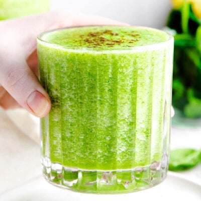 hand holding green smoothie in ridged glass with greens in the background.