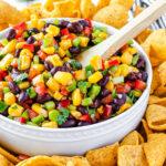 corn salsa with black beans in white bowl and wood spoon inserted. corn chips surrounding the bowl with limes in the background.