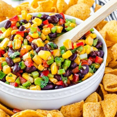 corn salsa with black beans in white bowl and wood spoon inserted. corn chips surrounding the bowl with limes in the background.