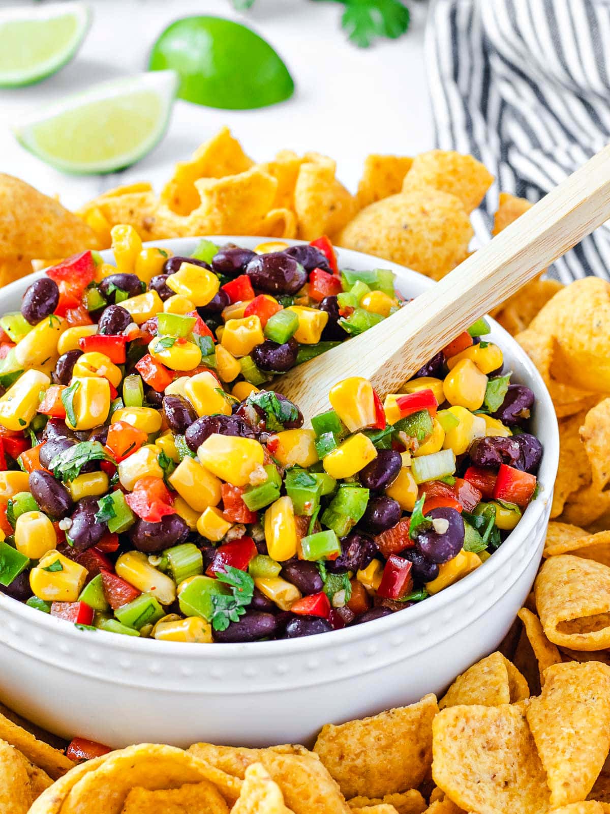corn salsa with black beans in white bowl and wood spoon inserted. corn chips surrounding the bowl with limes in the background.