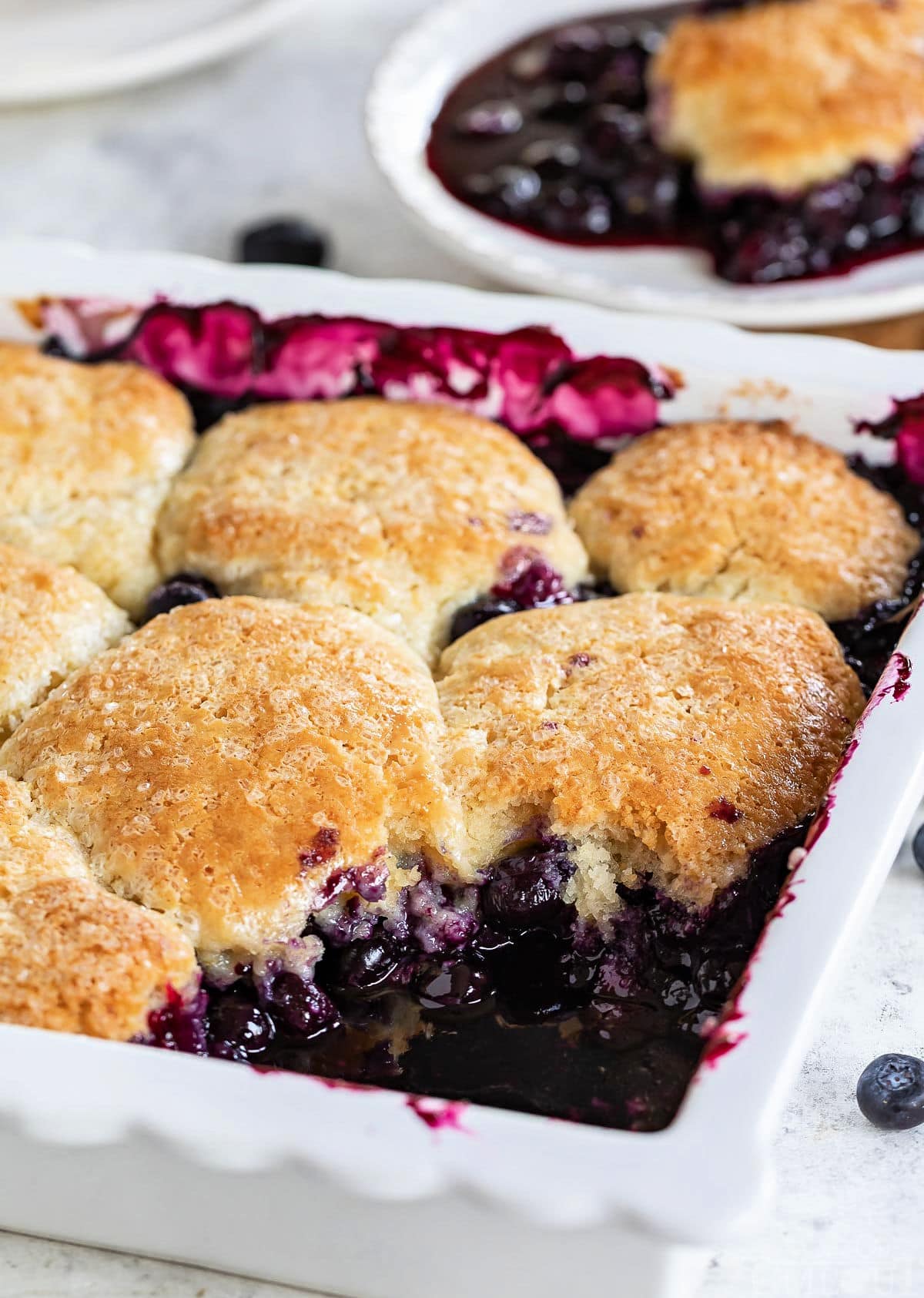 white baking dish with blueberry cobbler and one serving removed and sitting on plate in the background.