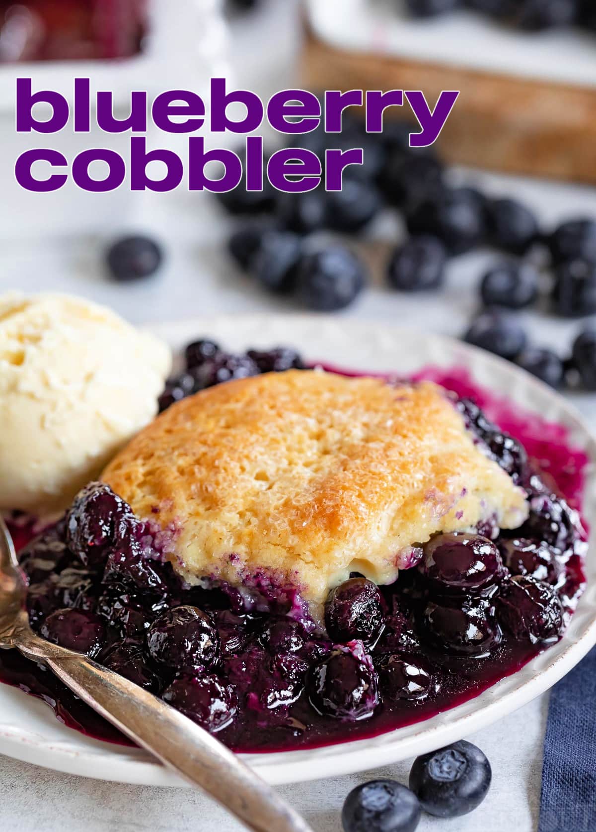 blueberry cobbler on small plate served with a scoop of vanilla ice cream. Fresh blueberries scattered about. Title overlay at top of image.