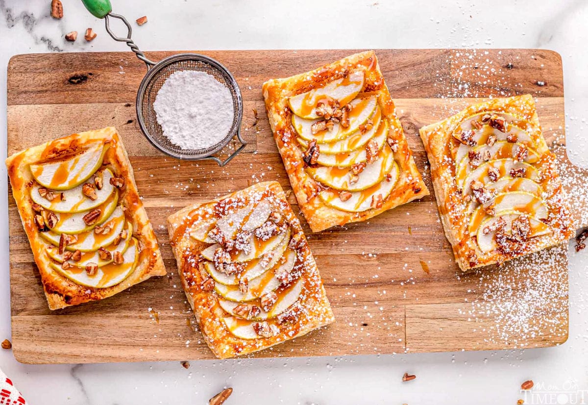 four apple tarts sitting on large wood cutting board drizzled with caramel and sprinkled with powdered sugar.