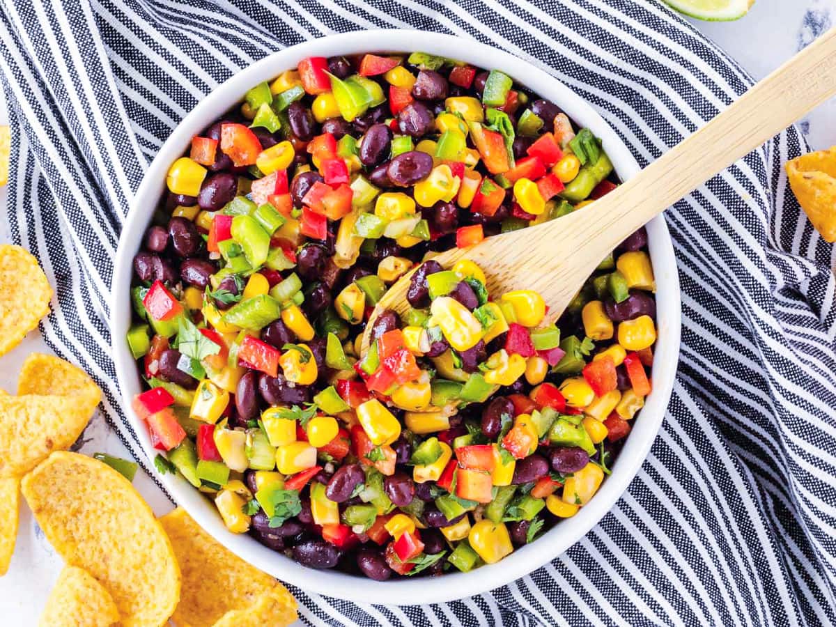 corn bean salsa in white bowl with wood serving spoon. bowl sitting on a black and white striped towel.