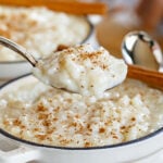 spoonful of rice pudding held up over the white pot it is being served from with freshly grated cinnamon stick on top.