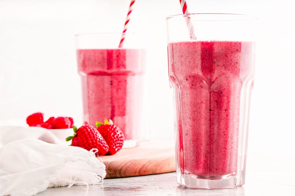 berry smoothie recipe in two tall clear glasses with paper straws. fresh strawberries and raspberries on a small wood cutting board next to the smoothies.