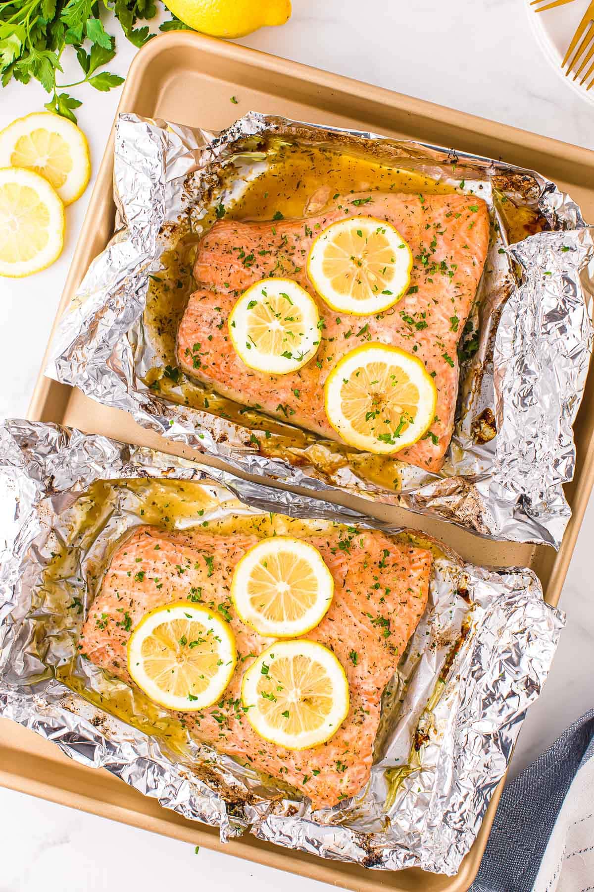 two large salmon filets baked in foil sitting on gold sheet pan topped with lemon slices and fresh minced parsley.