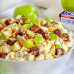 snickers salad in white salad bowl topped with a drizzle of caramel sauce. green apples and snickers candy bar in background.