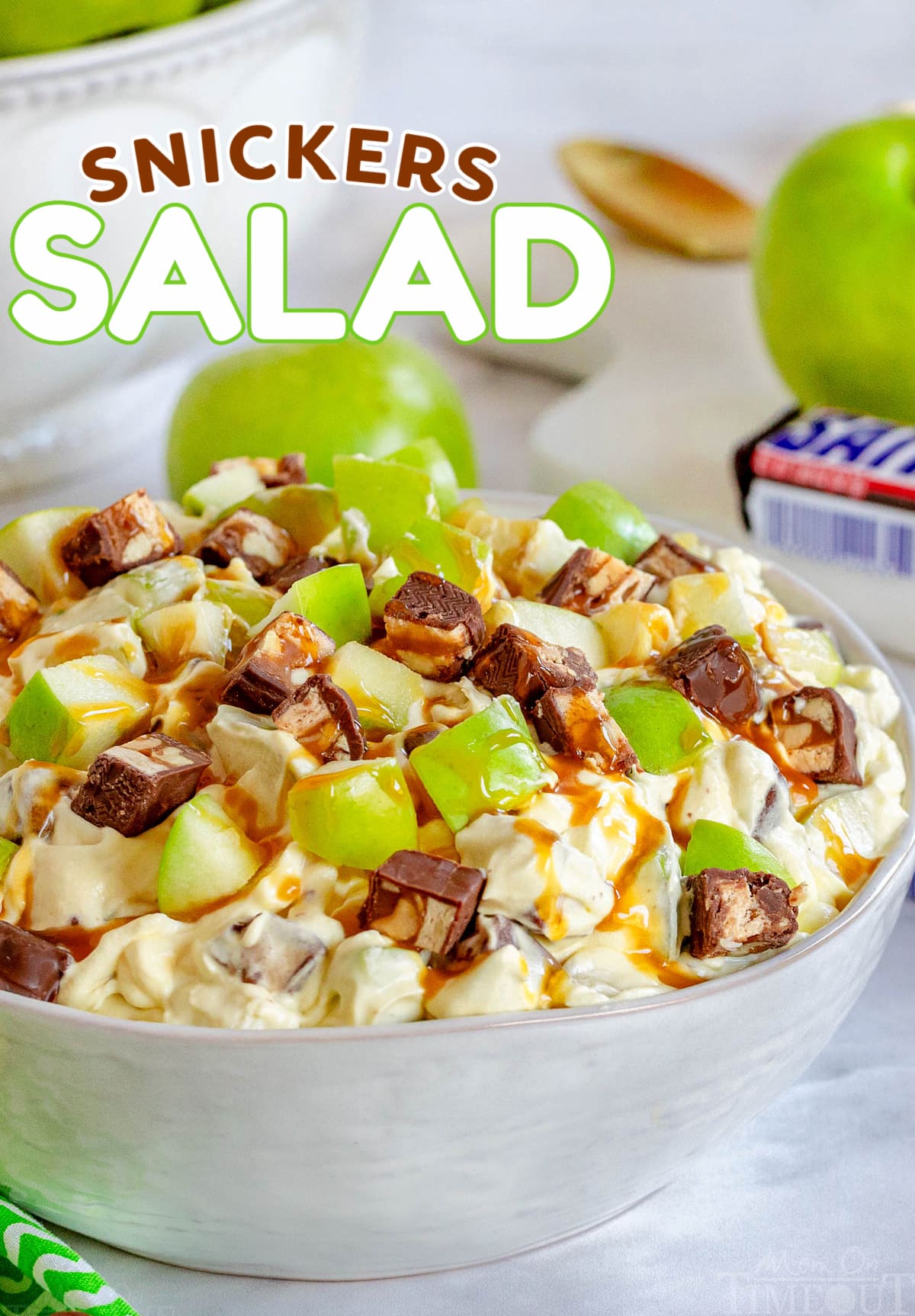 snickers salad in white salad bowl topped with a drizzle of caramel sauce. green apples and snickers candy bar in background. title overlay at top of image.