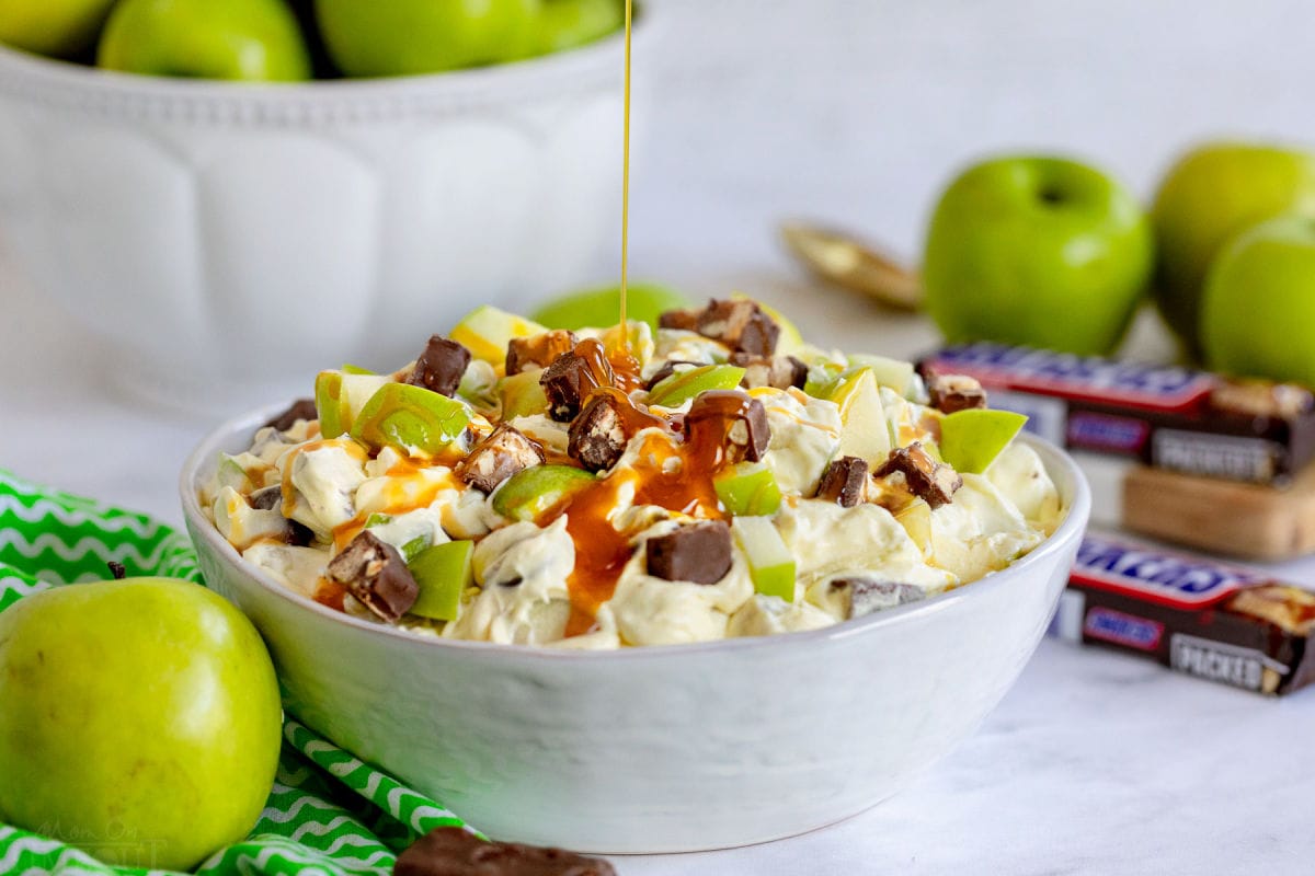 caramel sauce being drizzled over the top of the snickers salad in large white bowl. apples and snickers around the bowl.