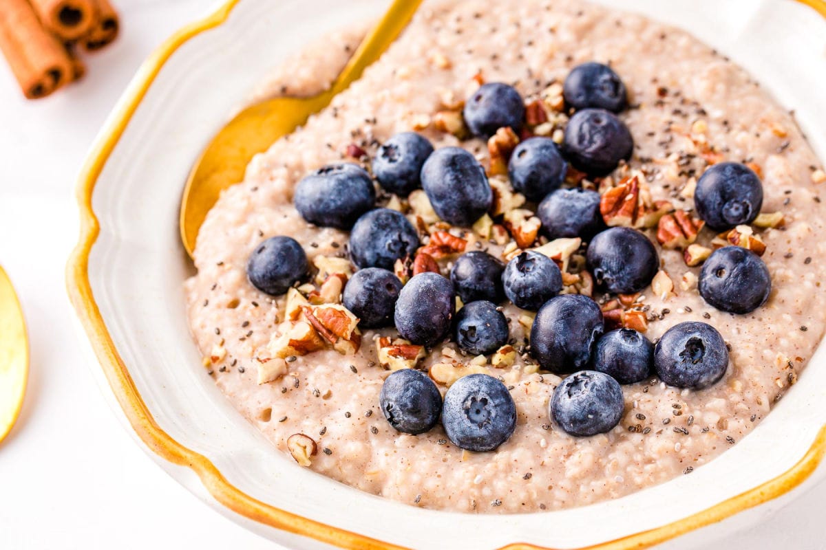 steel cut oatmeal topped with blueberries, pecans and cinnamon in a white bowl with a gold spoon.