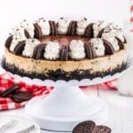 whole cheesecake made with oreos on a white metal cake stand and topped with whipped cream and oreo cookies.