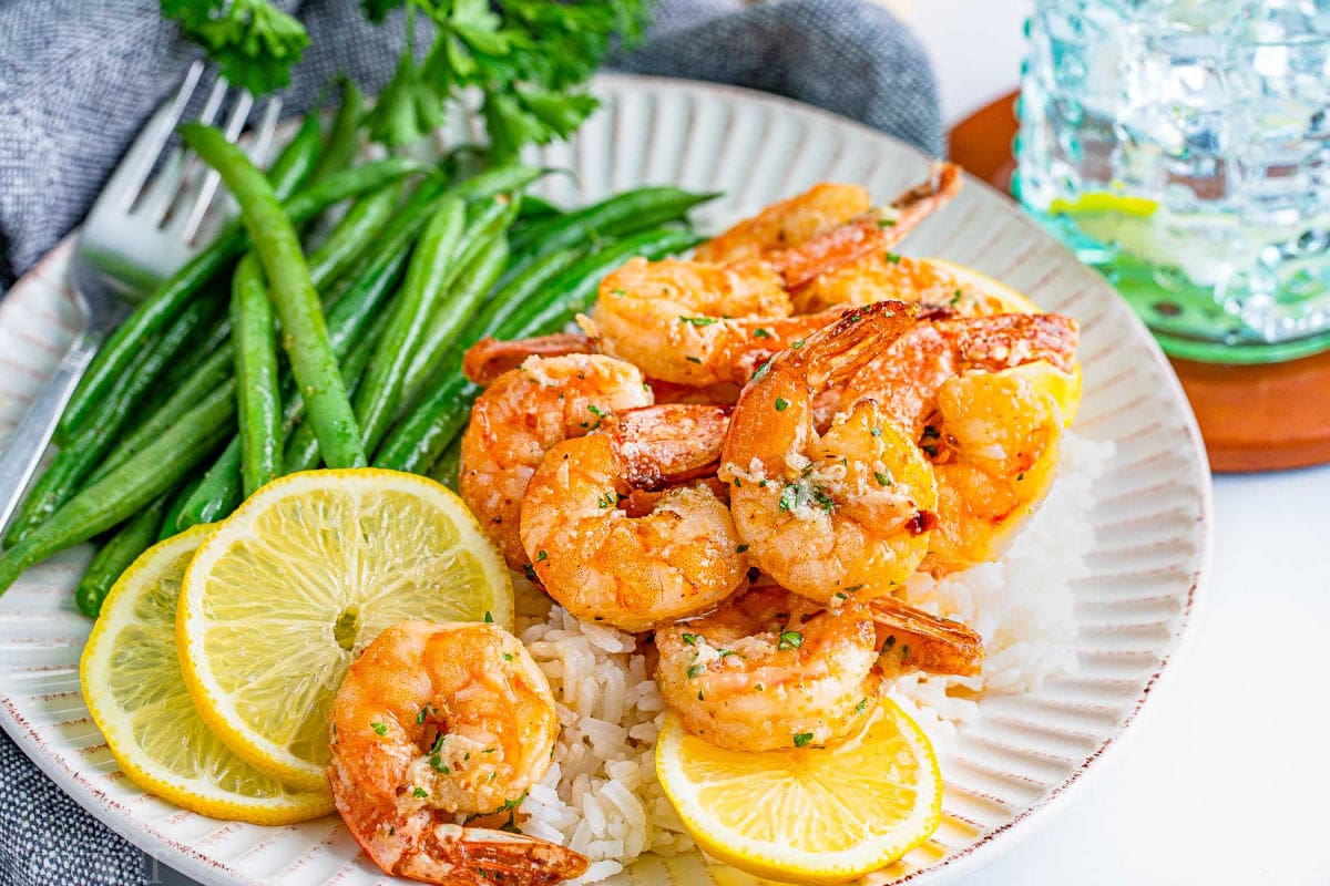 air fryer shrimp with garlic her butter sauce sitting on white scalloped edge plate with green beans and lemon slices.