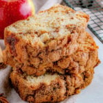 three slices of apple bread stacked next to a cooling rack with an whole red apple in background.