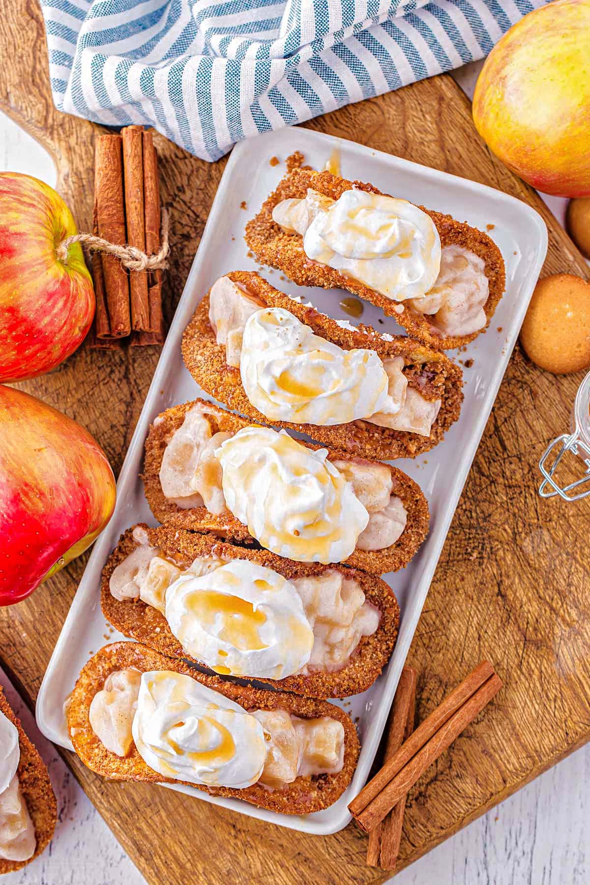 top down view of dessert tacos filled with apple pie filling and topped with whipped cream and caramel all lined up on a white tray ready to serve.