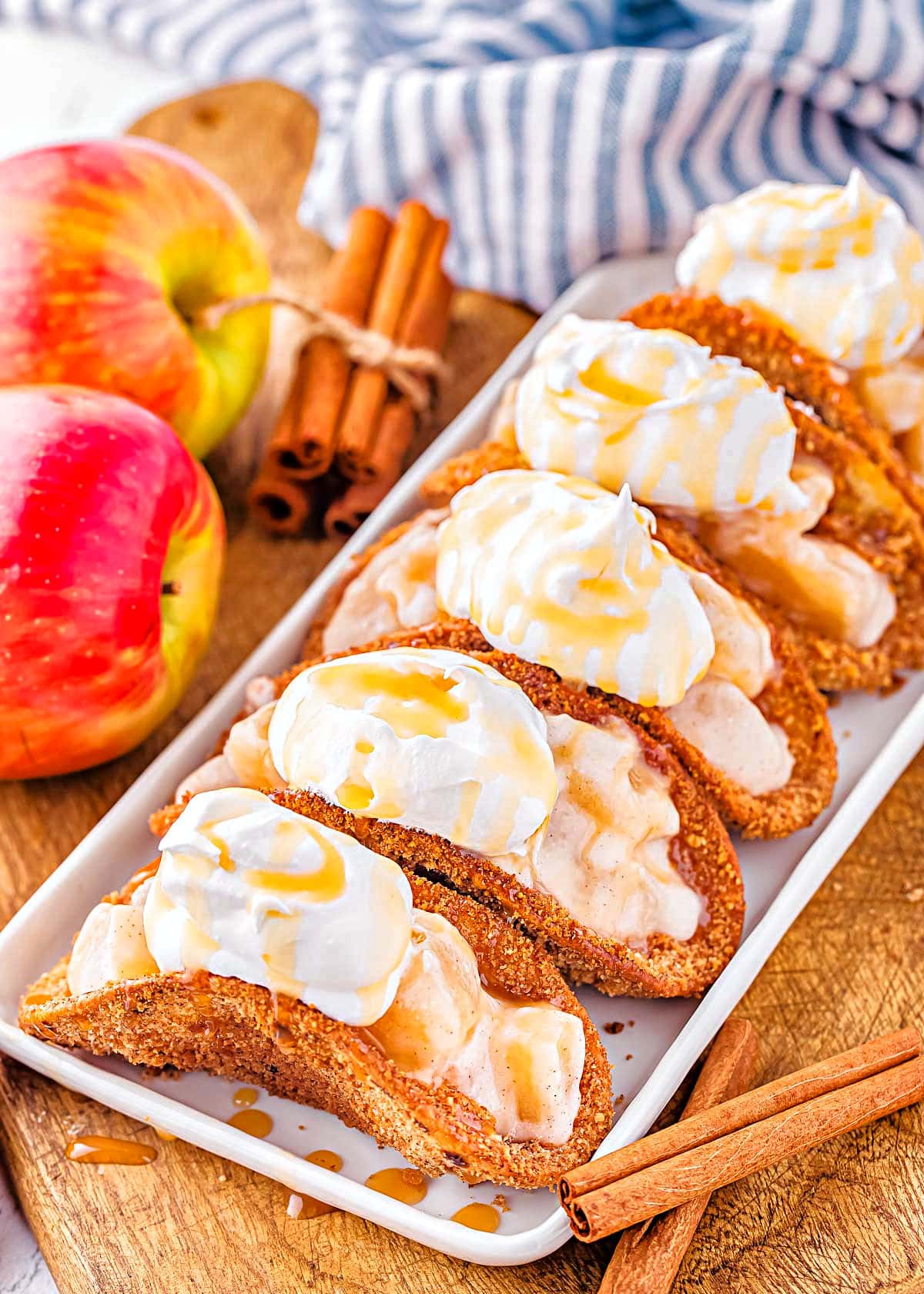 5 apple pie tacos on shallow white serving dish sitting next to blue and white striped napkin and fresh red apples.