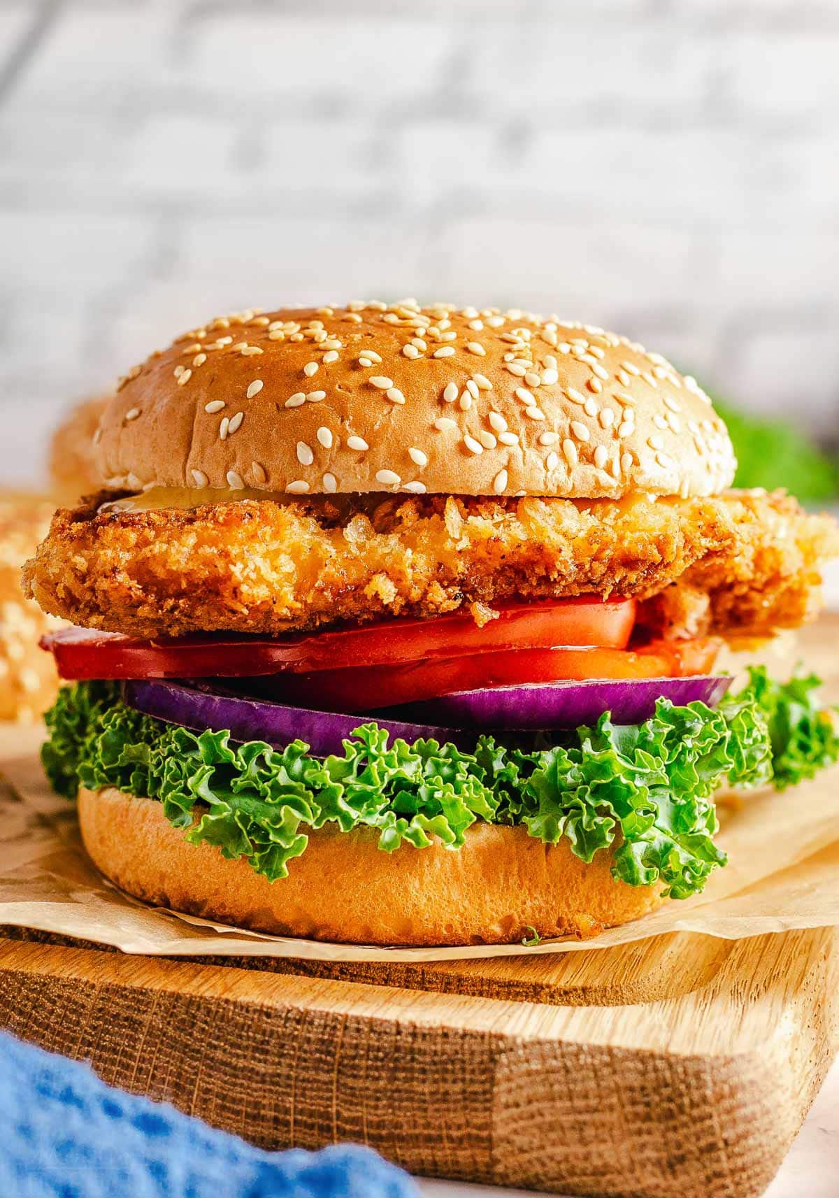 crispy chicken sandwich with all the topping on a seeded hamburger bun sitting on brown parchment on a wood cutting board.