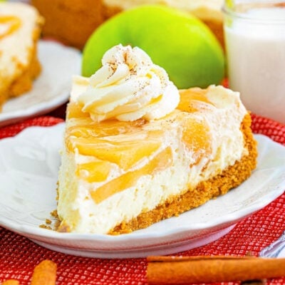 slice of apple cheesecake sitting on a white plate with fork. cheesecake is garnished with whipped cream and apple pie spice. whole cheesecake in background.