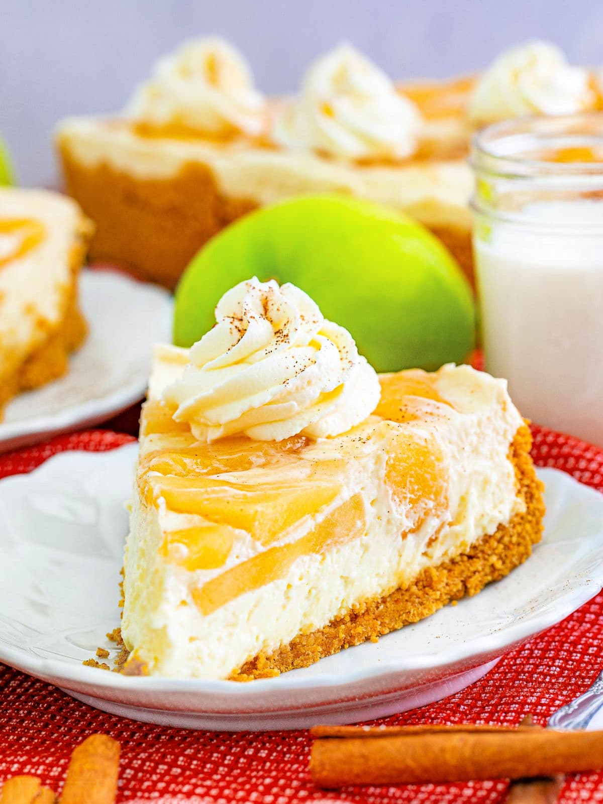 slice of apple cheesecake sitting on a white plate with fork. cheesecake is garnished with whipped cream and apple pie spice. whole cheesecake in background.