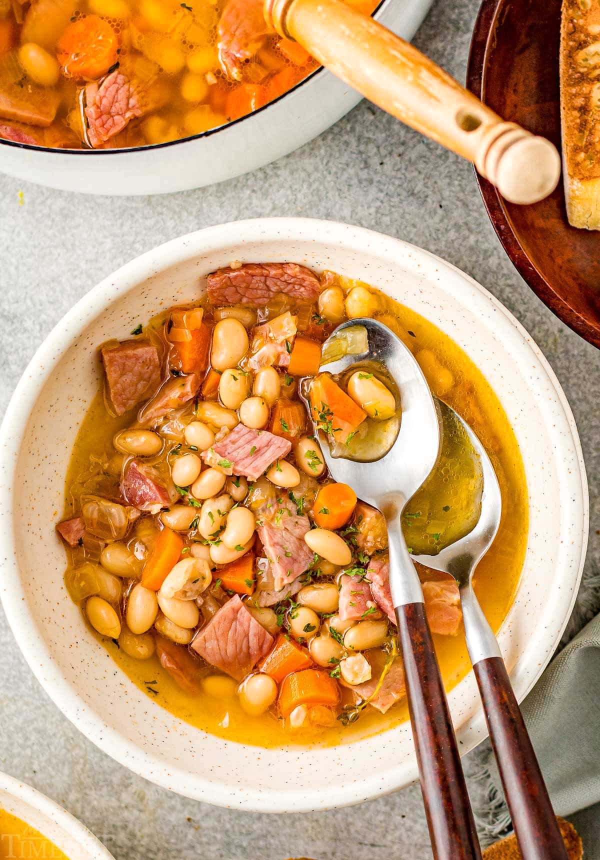 bowl of ham and bean soup with two spoons with wood handles next to pot of soup.