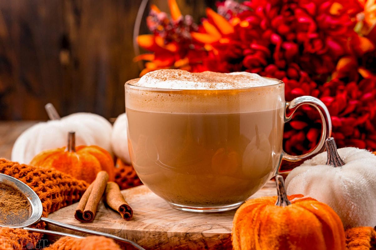 front view of a pumpkin spice latte ready to be enjoyed garnished with pumpkin pie spice and sitting in front of fall colored flowers on a wood board.