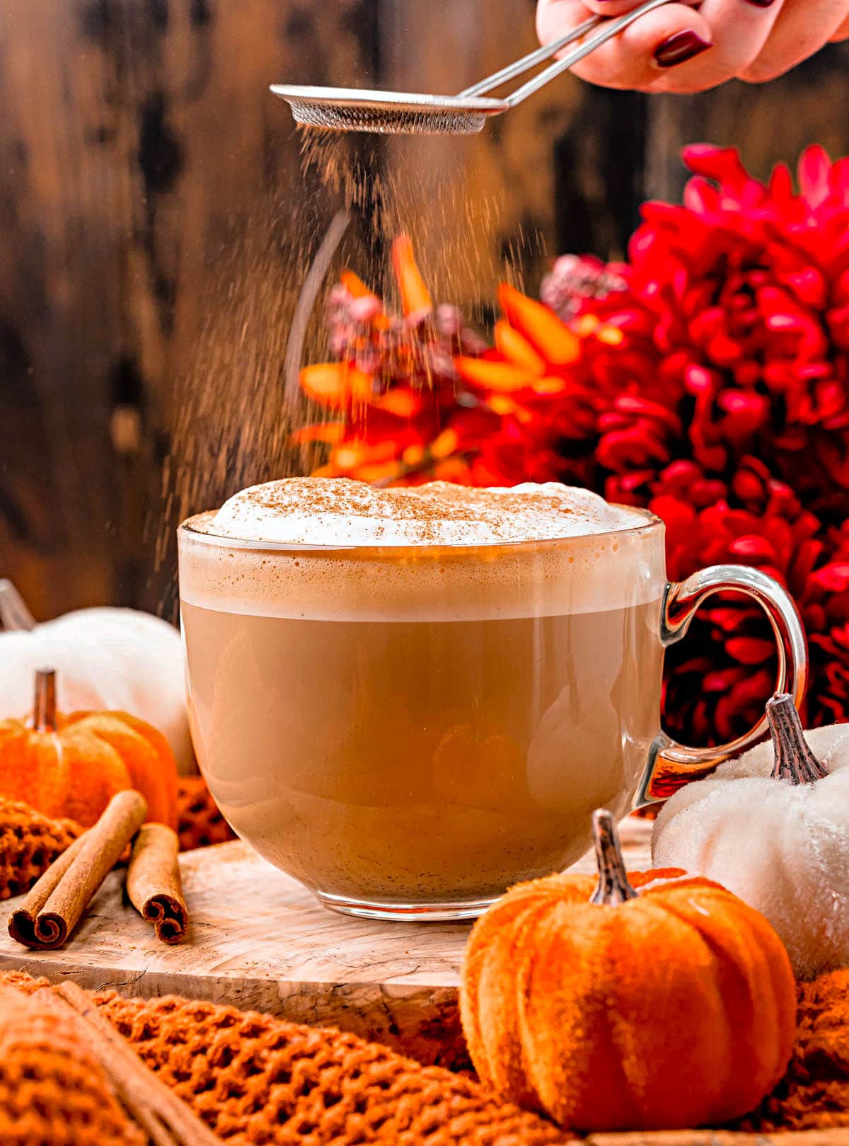 pumpkin spice latte in glass mug with pumpkin spice being dusted on top of the foam topping. colorful fall flowers in background and small velvet pumpkins scattered about.