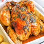 roasted chicken with golden skin and herbs cooked on top sitting in white roasting dish.